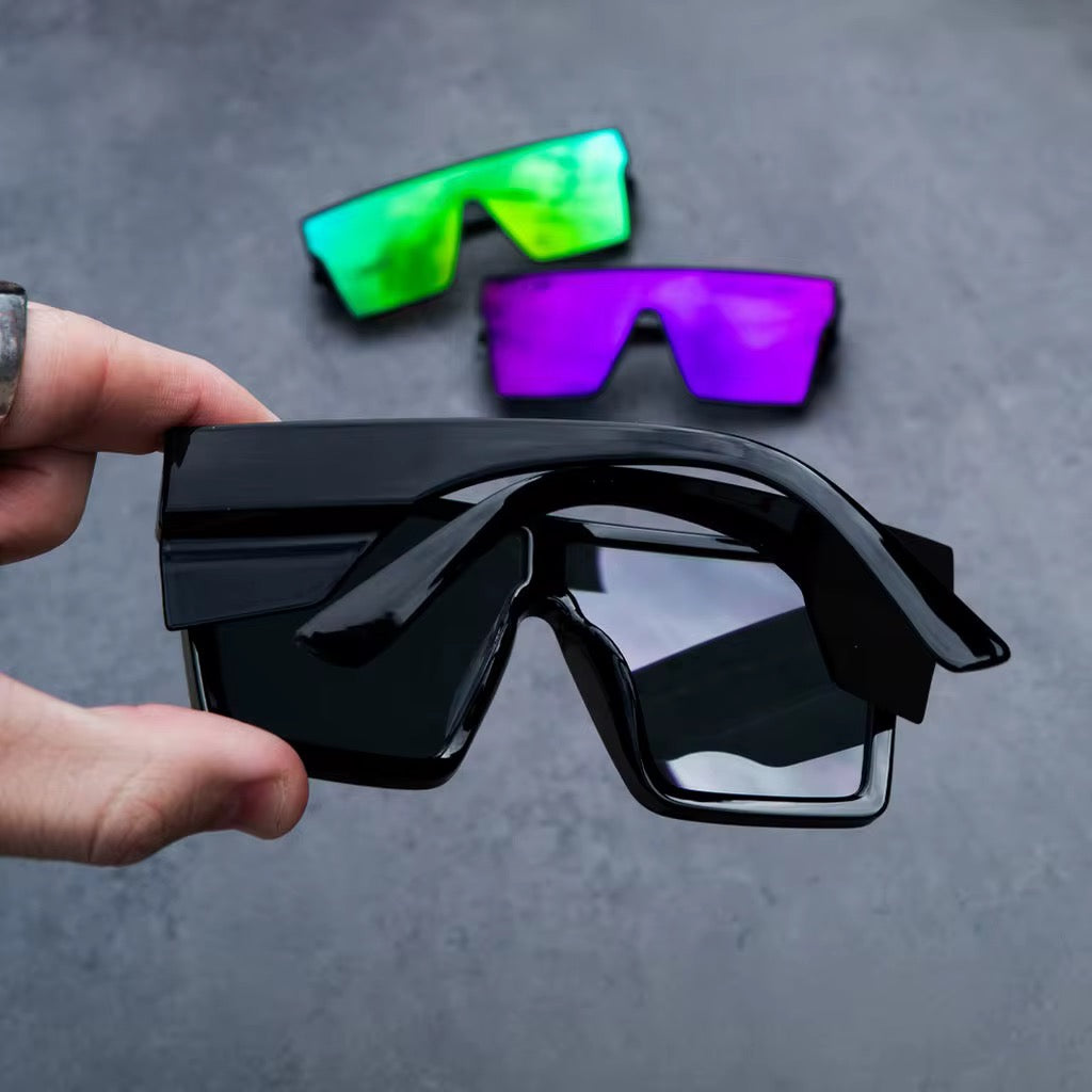 Hand holding stylish black sunglasses with colorful shades in the background on a gray surface.